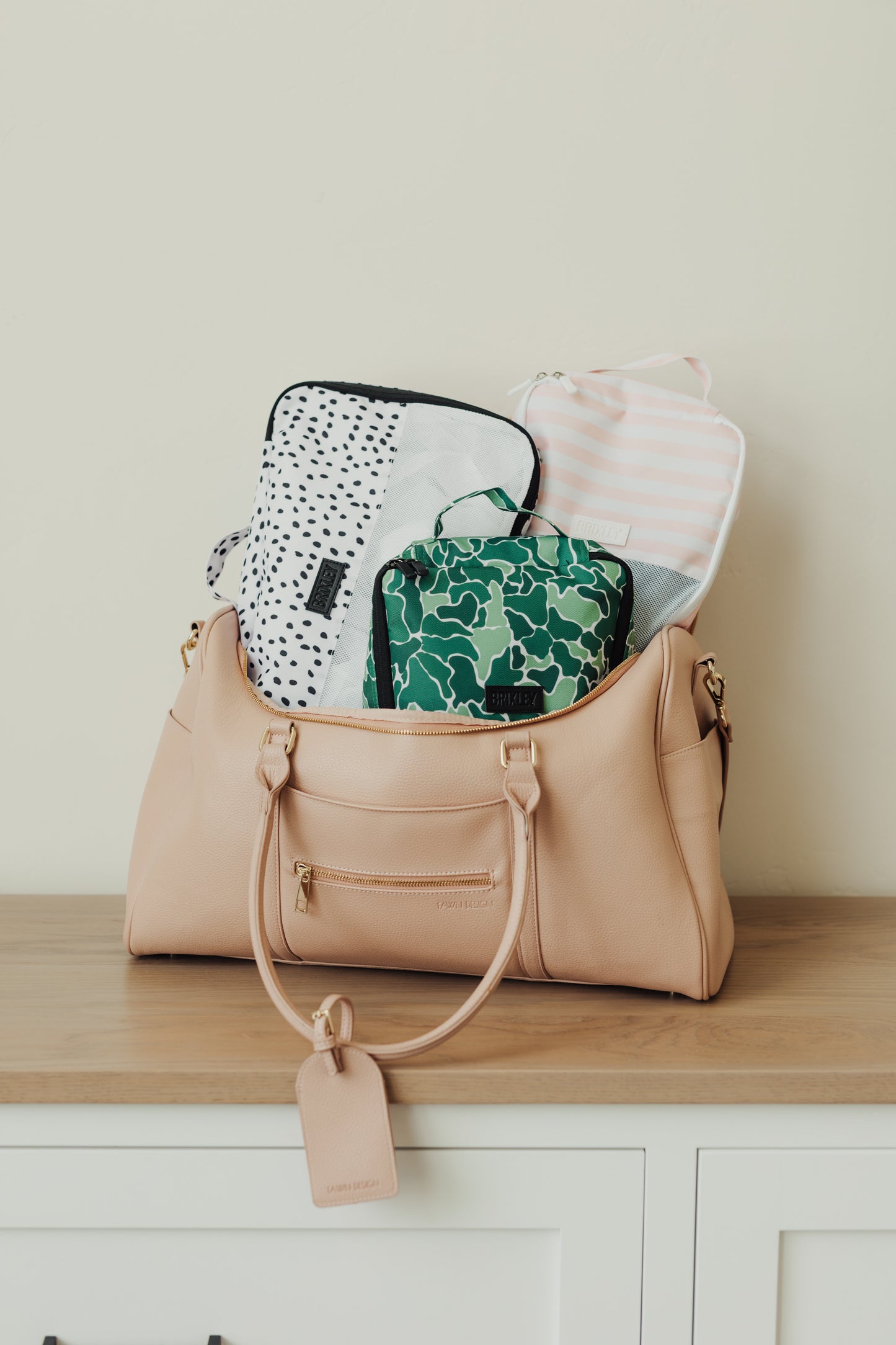 Three (3) Brixley Bag Packing cubes packed into a tan bag. One (1) Dalmatian print, one (1) Camouflage (Scout) print, one (1) white and pink striped (Valentine). All sitting on top of a wood countertop 